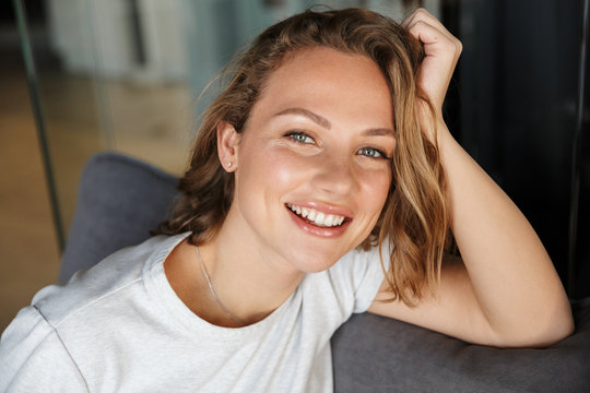 Image of happy caucasian woman smiling at camera while sitting on sofa