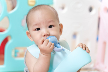 An Asian baby sits in a pen, biting a pacifier