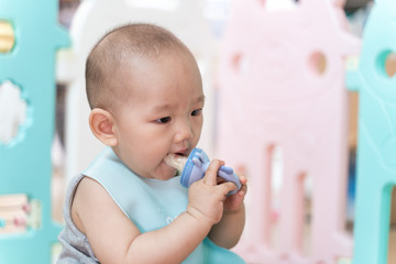 An Asian baby sits in a pen, biting a pacifier