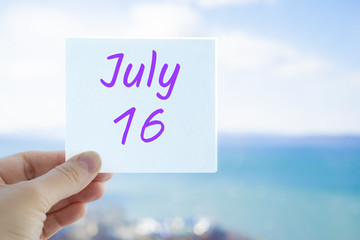 July 16th. Hand holding sticker with text July 16 on the blurred background of the sea and sky. Copy space for text. Month in calendar concept