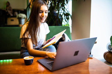 Obraz premium Young woman sitting at table and taking notes in notebook with table is laptop, smartphone and cup of coffee.