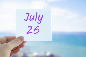 July 26th. Hand holding sticker with text July 26 on the blurred background of the sea and sky. Copy space for text. Month in calendar concept