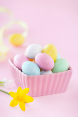 Beautiful pastel pink Easter composition - Colourful candy Easter eggs in a bowl with a daffodil at the foreground against pink background