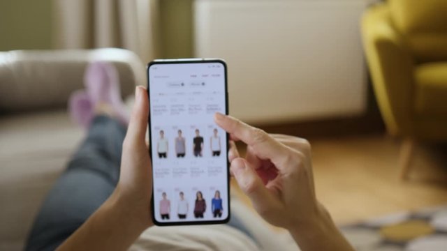 Woman Looks at Goods in Online Store Clothing. Buy Fashion Clothes Directly on Smartphone. Woman at Home Lying on Couch in Living Room Using Smartphone. Screen is blurred. Focus on hand.