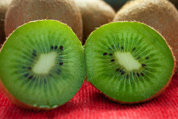 One kiwi cut in half close up on a red surface. In the background others kiwis. 