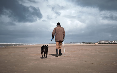A retired old man with a walking stick walking his dog on a beach. Dogs are mans best friend and a loyal companion for older people who are alone. Love animals and pets.