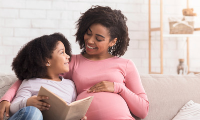 Little black girl and her pregnant mom enjoying reading book together