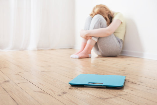 Sad Teenager Girl With Scale On Wooden Floor