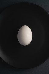 Single white egg in black plate on dark moody plain minimal background, top view, happy Easter day