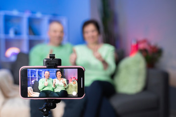 Elderly couple using smartphone video conference with grandchild while lying on sofa in living room at home. Enjoying time lifestyle senior family at home concept. Portrait looking at camera.