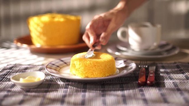 Female hand butters yellow couscous on a plate on a table with a white porcelain cup in the background.