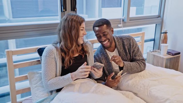 Young multiracial couple sitting in bed, scrolling through social media and laughing at photos/videos on their smartphones