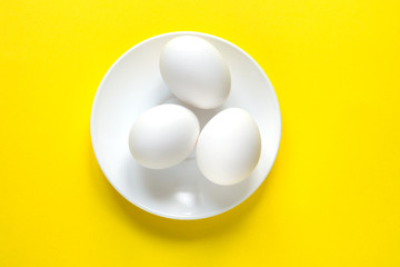 Three white eggs on a round plate lie on a colorful yellow background.