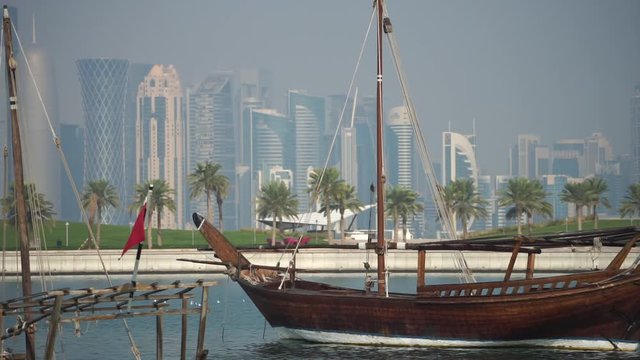 Retro historical boat with blurred panoramic view of modern skyline of Doha and green palms on background