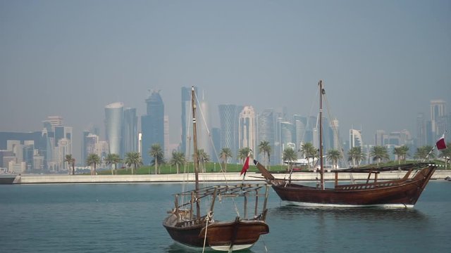 Retro historical boat with blurred panoramic view of modern skyline of Doha and green palms on background