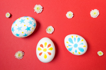 Easter concept. White eggs with marker drawings of petals and stars. Manual coloring. Yellow, red and blue shades. Red background. Nearby are small white flowers. Flat lay, top view, copyspace.