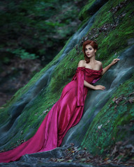 Fototapeta premium Stunningly beautiful cute young woman with fiery red hair in long red dress with train stands in green forest leaning on stone green slope and looking at camera