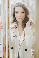 Through window portrait of young fashionable woman in coat posing to camera while standing on home terrace. Beautiful affectionate girl in stylish clothing posing on balcony. Gentle fashion model