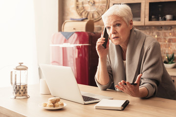 Concerned elderly woman talking on cellphone, working on laptop in kitchen