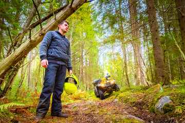 The man next to the ATV looks around. Biker is resting. A man came to the forest on a quad bike. Concept - a journey through the forest. Guy holds a biker helmet in his hands. Travel on the off-road