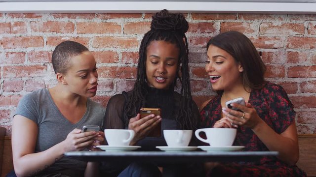 Three female friends meeting for coffee sitting at table looking at mobile phones together - shot in slow motion