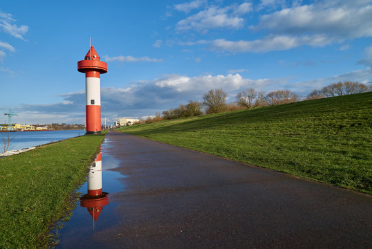 small lighthouse at Ritzenb&uuml;tteler Sand, municipality Lemwerder (district Wesermarsch, Germany) on a sunny spring day with vivid blue sky and clouds reflecting in a puddle on the footpath next to dike