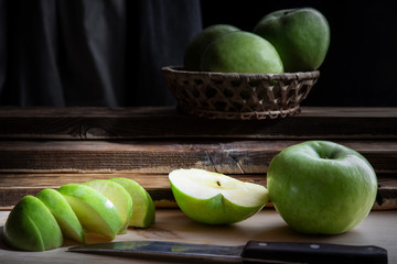 Still life of green apples lying in a basket and on boards, on a black background with light illumination, shallow depth of field. Conception, healthy food.
