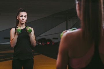 Back view of young sports woman looking at her reflection in mirror while holding dumbbells in hands. Athletic beautiful girl training with dumbbells against mirror. Exercising and workout in dark gym