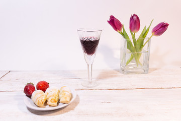 Vintage glass of red wine, raspberries and croissants on plate, pink tulips in vase on white background