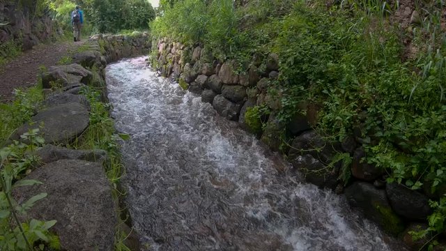 Tourists walking along a path surrounded by a lot of vegetation next to a clear water stream in the sacred valley of the incas, located in Cusco, Per&uacute;.