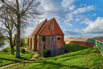 Plakat small brick church in Warfleth (municipality Berne, district Wesermarsch, Germany) built into the dike on a sunny spring day with vivid blue sky and clouds