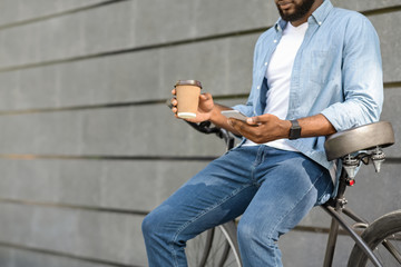 Fototapeta na wymiar Black millennial guy with smarthone and takeaway coffee sitting on bicycle