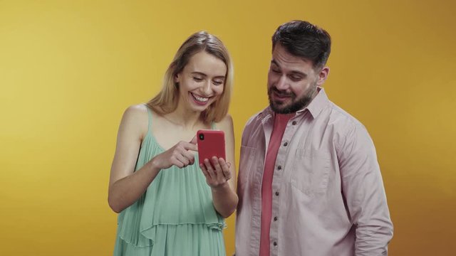 Romantic caucasian couple of man and woman friends using smartphone together laughing at funny content compilation posing at orange background.