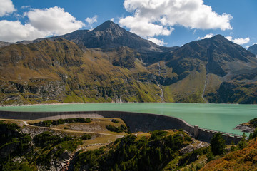 A view of the Alpine dam at the foot of the rocky peaks under the blue sky with nice clouds