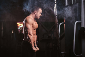 Fototapeta na wymiar Strong shirtless male athlete concentrated on an excersise on a hand pull machine, pumping up his triceps in a dark gym surrounded by smoke