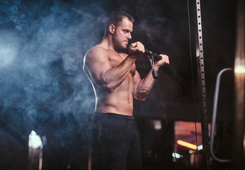 Powerful adult bodybuilder pumping up his biceps on a hand pull machine in a dark gym under the spotlights surrounded by smoke looking serious