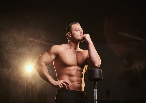 Fit adult man with ideal body leaning on the barbell in a thinking pose in a dark gym surrounded by smoke