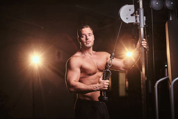 Fototapeta na wymiar Athletic shirtless sportsman doing an excersise on a hand pull machine in a dark gym under the spotlights surrounded by smoke