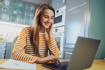 Happy young freelancer woman working at home using modern laptop device, cheerful student girl in casual clothes with long hair browsing internet studying with laptop computer at cozy home interior 