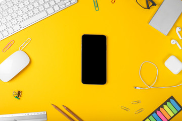 Mockup of smartphone on the background of office items on yellow background. Computer mouse and keyboard, pencils, headphones, smart watch and eye glasses. Top view with copy space, flat lay.