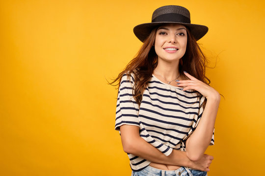 A Beautiful Brunette In A Striped T-shirt And Wicker Hat Stands On A Yellow Background, Smotot Into The Frame,raising Hand To Neck