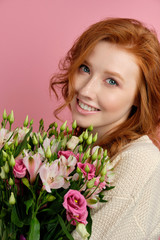 Obraz na płótnie Canvas A red-haired curly girl in a white sweater stands on a pink background clutching a bouquet of flowers and looks into the frame