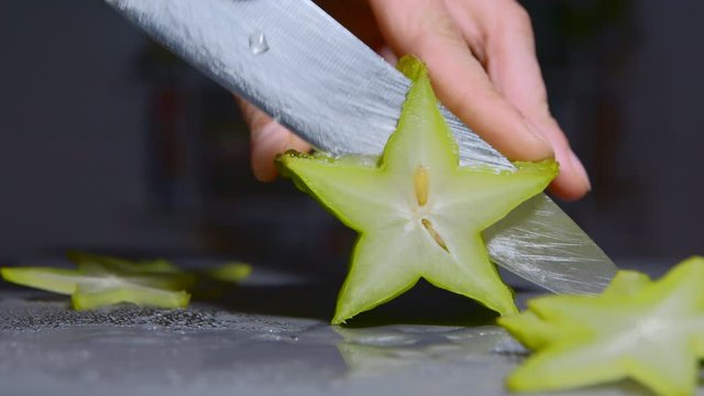 Cutting starfruit with seeds into pieces. Cooking dessert, sharp knife cuts carambola. The play of light and shadow. Dark background, close-up. Macro 4K