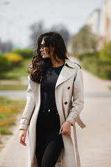 Seductive young woman with perfect body in beige raincoat and black jeans posing on the city street on the sunny spring day. Brunette model girl in fashionable spring outfit posing at urban background