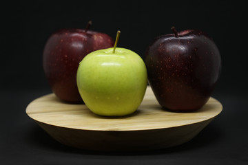 Green and two dark red apples on a wooden plate isolated on a black background.