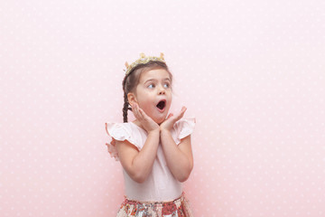 Surprised pretty young girl in pink dress with crown on head expressing isolated on pink...