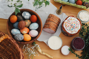 Easter food flat lay. Easter eggs natural dyed, easter bread, ham, beets, butter, green branches  and flowers on rustic wooden table with wicker basket and candle. Traditional Easter Food