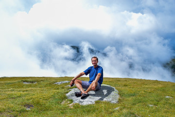 Fototapeta na wymiar The man enjoys the sun's rays, sitting on a stone surrounded by fog and low clouds near Transalpina road. This is one of most beautiful alpine routes and highest mountain asphalt road in Romania.