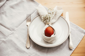 Stylish Easter brunch table setting with egg in easter bunny napkin on  table. Natural dyed easter egg on napkin with bunny ears with flowers on plate and cutlery. Easter table decorations