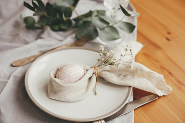 Fototapeta na wymiar Stylish Easter brunch table setting with egg in easter bunny napkin on table. Modern natural dyed pink easter egg on napkin with bunny ears, flowers on plate and cutlery. Easter table decorations.